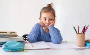menininha sentada com caderno estojo e lapis sobre a mesa com expressao de falta de atencao