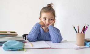 menininha sentada com caderno estojo e lapis sobre a mesa com expressao de falta de atencao