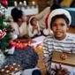 menino com gorro de natal sentado no chao segurando caixa de presete com arvore de natal e familia ao fundo