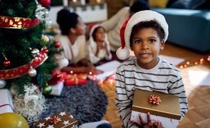 menino com gorro de natal sentado no chao segurando caixa de presete com arvore de natal e familia ao fundo